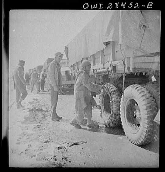 Somewhere in the Persian corridor, a United States Army truck convoy carrying supplies for the aid of Russia. An Iranian boy giving the American driver a helping hand at putting on chains before the trucks go into a snow bound pass through the mountains.