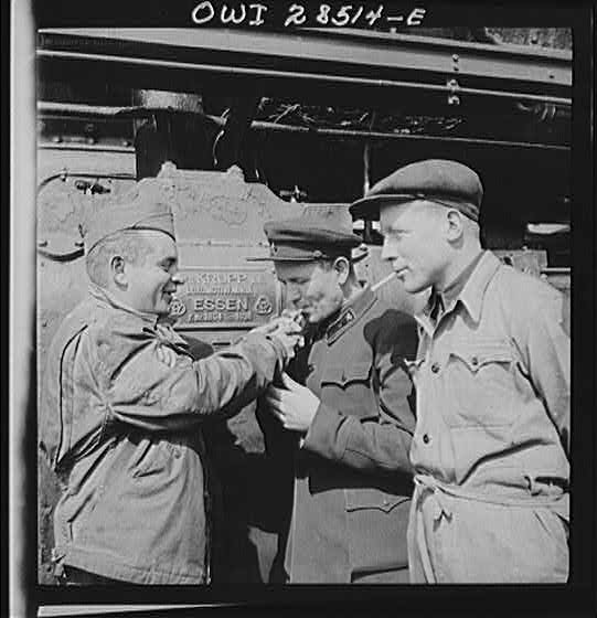 An American Sergeant railroader lighting the cigarette of two Russian railroad engineers as they stand before a German-made engine being used to haul supplies for a Russian railway. Sign on the engine shows that it was built at the Krupp works in Essen. Somewhere in Iran.