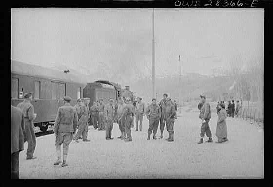 American and British soldier trainmen standing about at a station on the route for supplies to Russia. An American engine is seen at the head of the train at left, somewhere in Iran.